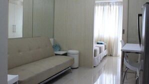 A modern hotel room with a sofa, a small table, a mirror, a refrigerator, and a window with curtains.