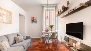 Una sala de estar moderna con un sofá gris, una mesa de comedor con sillas, un televisor de pantalla plana en la pared y piso de madera.