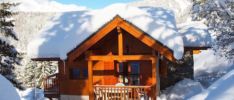A wooden cabin with a porch, surrounded by snow-covered trees and mountains.