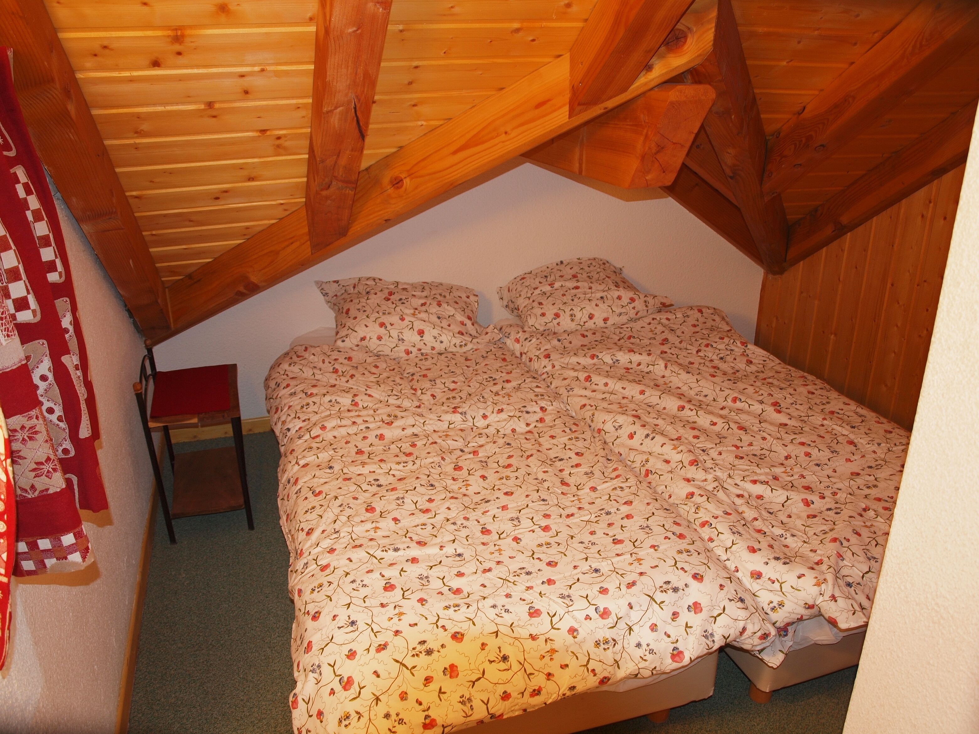 Une chambre avec un plafond en bois, un lit recouvert d’une literie à motifs floraux et une petite table de chevet.