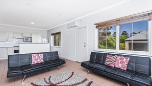 A modern living room with two black leather sofas, a kitchen area with white cabinets, and large windows offering a view of greenery. - Bella On Victoria - with ocean views (Port Fairy)