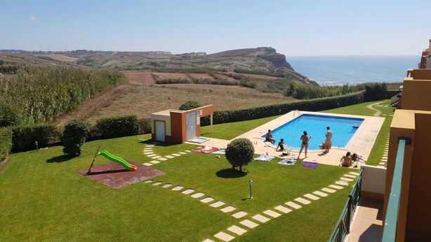A swimming pool with a view of the sea, a small play area, and a grassy lawn.