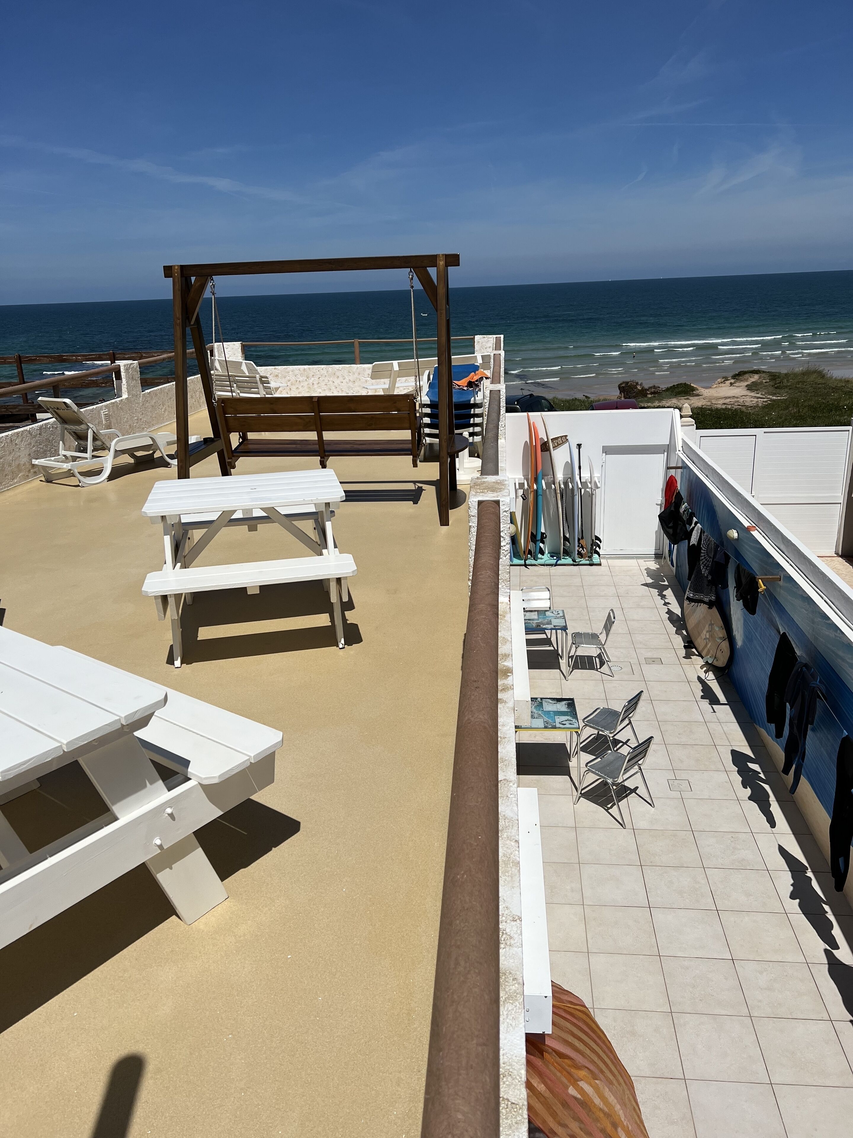 A beachside patio with white tables and benches, a wooden swing, and a surfboard rack.