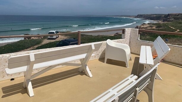 Blick auf einen Strand mit einer weißen Bank und einem weißen Stuhl, einem Sandbereich und einem Auto in der Nähe des Wassers.