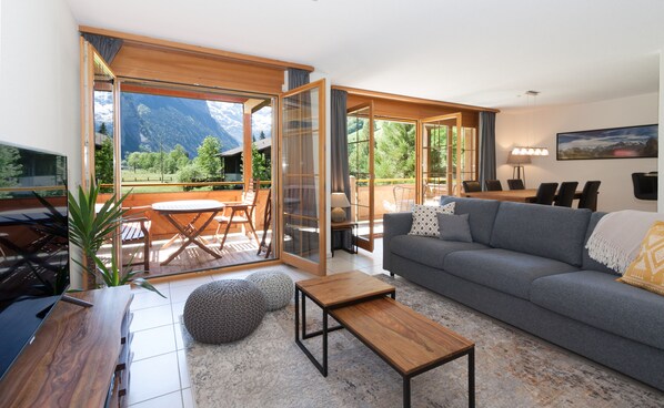 A modern living room with a grey sofa, wooden coffee table, and a dining area with a table and chairs. Large glass doors lead to a balcony with a mountain view. - Apartment Arven - 3 Bedroom - 2 Bathroom - Uninterrupted mountain view (Lauterbrunnen)
