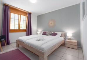 A bedroom with a wooden bed, purple curtains, a wall clock, and a bedside table with a lamp. - Apartment Arven - 3 Bedroom - 2 Bathroom - Uninterrupted mountain view (Lauterbrunnen)