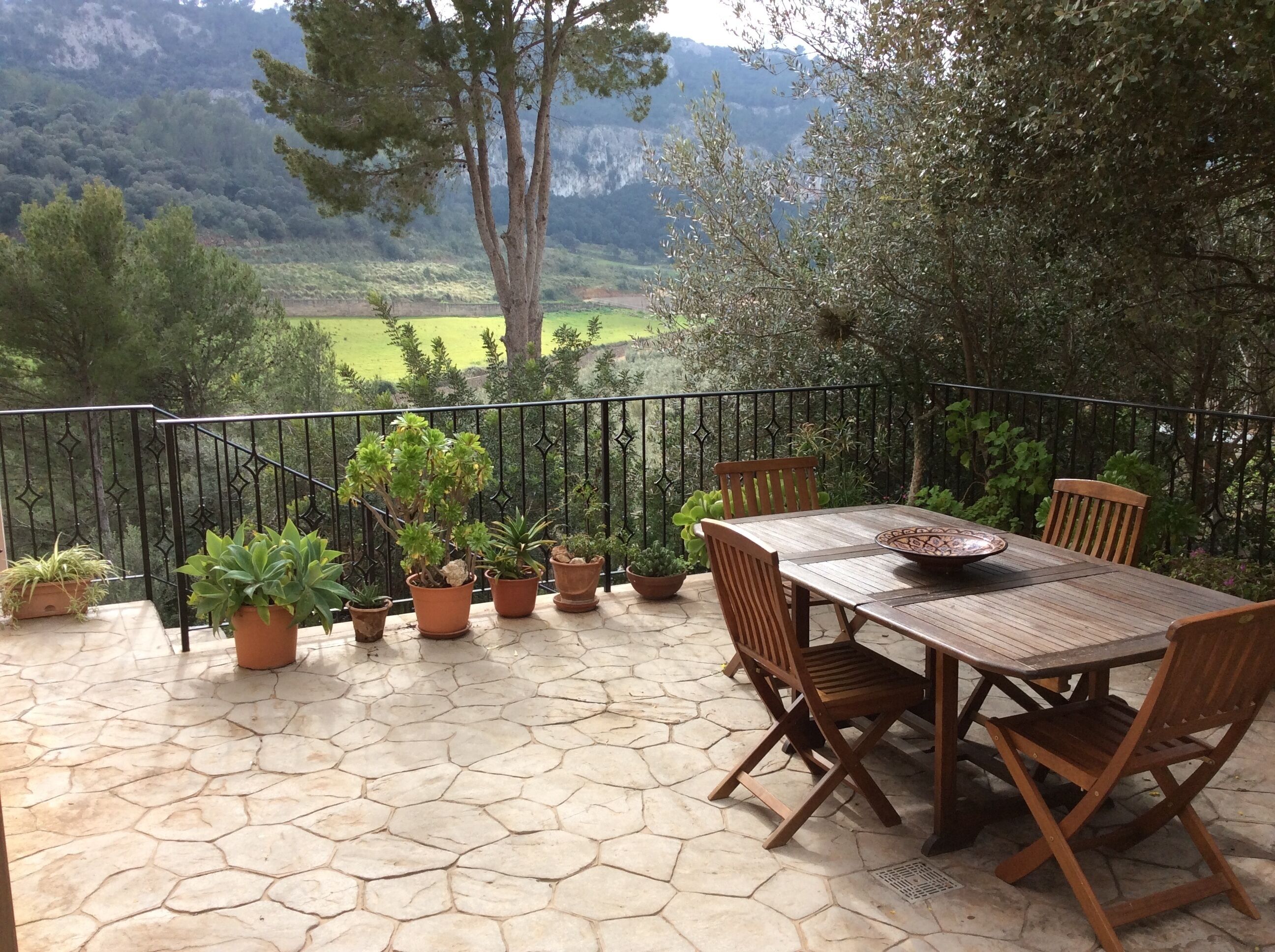 A patio with a table and chairs, surrounded by potted plants and a view of a mountainous landscape.
