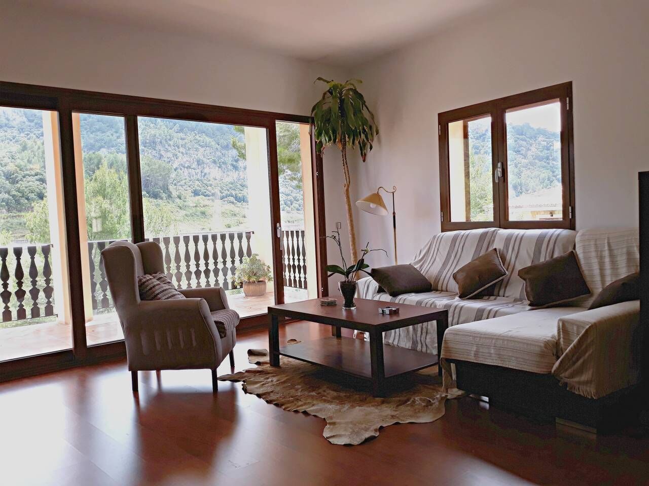 A spacious living room with a sofa, armchair, and coffee table, large windows with a view of a mountainous landscape, and a potted plant.