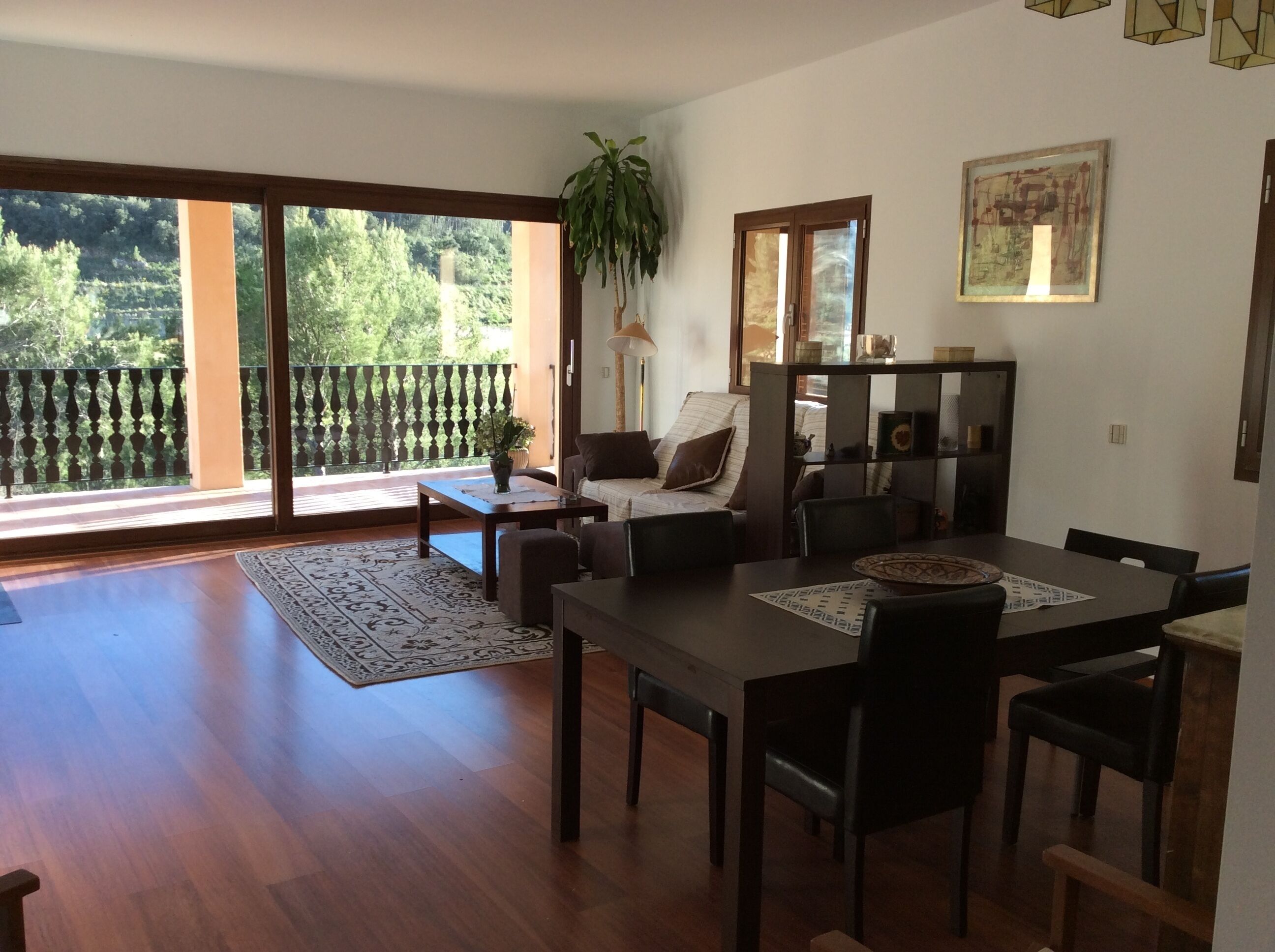 A spacious living room with a wooden floor, a dining table with chairs, a sofa, and a large sliding glass door leading to a balcony.
