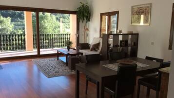 A spacious living room with a wooden floor, a dining table with chairs, a sofa, and a large sliding glass door leading to a balcony.