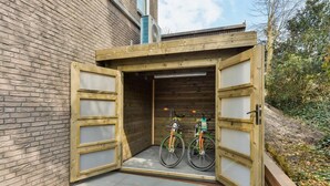 Un cobertizo de madera para bicicletas con dos bicicletas en su interior, junto a un edificio de ladrillo.