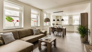 Una sala moderna con sofá, mesa de centro y zona de comedor con sillas. Hay ventanas amplias con flores y una cocina al fondo.