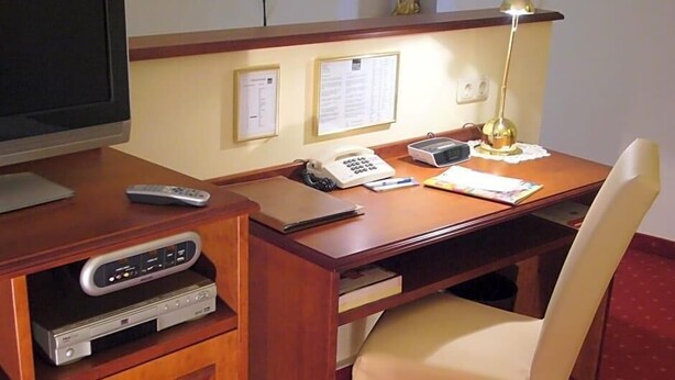A hotel room with a wooden desk, a television, a remote control, a telephone, a lamp, and a chair. - Klassik Hotel Am Tor (Weiden in der Oberpfalz)