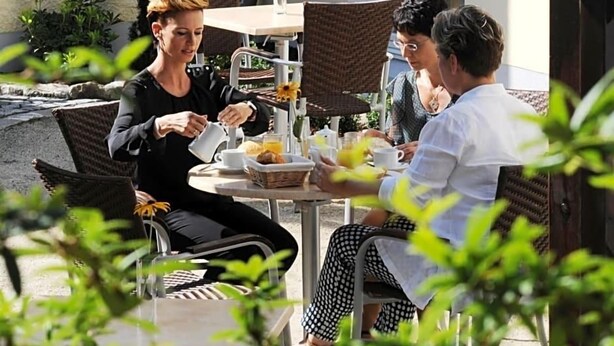 A group of people dining outdoors at a table with wicker chairs, a basket of bread, and a cup of coffee.