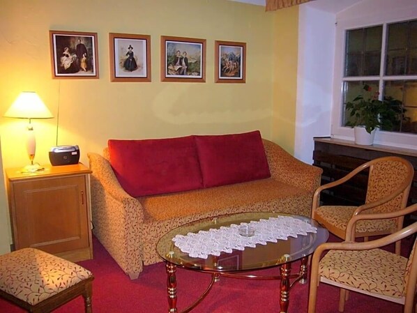 A living room with a sofa, a glass coffee table with lace, a lamp, a chair, and framed pictures on the wall.