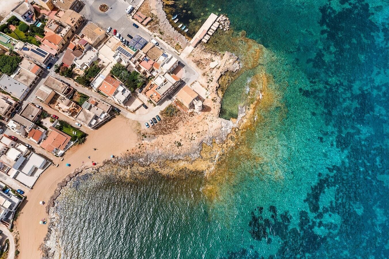 Vista aérea de uma área costeira com edifícios, praia e águas azul-claras.
