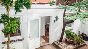Een wit gebouw van baksteen met een open deur naar een kamer met een open haard, een klein tuintje met een boom en een bloempot.