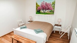 Een slaapkamer met een wit bed, een houten bankje, twee witte stoelen, een witte nachtkastje met een lamp, en een schilderij van een boom aan de muur.