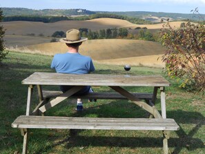 Une personne est assise à une table de pique-nique en bois, un verre de vin à la main, et contemple des collines ondoyantes ainsi qu’un bâtiment situé au loin.