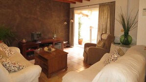 Una sala de estar con una mesa de centro de madera, un sofá, una silla y un televisor sobre un mueble de madera. Hay una planta en maceta en el alféizar de la ventana y un jarrón con plantas verdes altas sobre una mesita de noche.