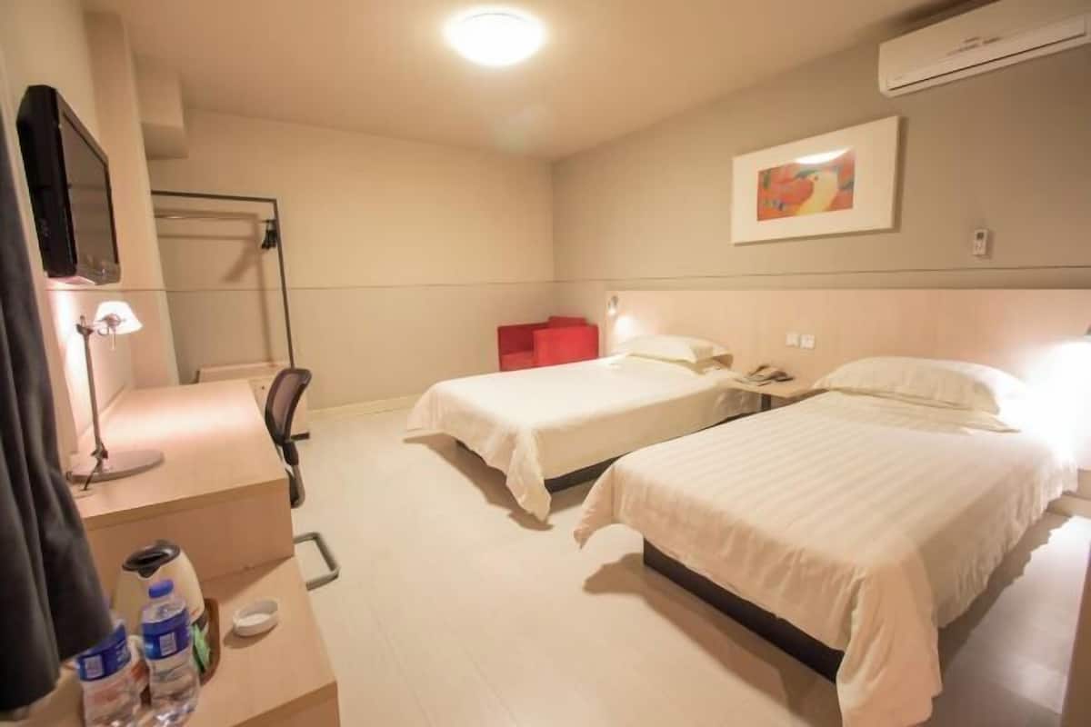 A hotel room with two beds, a desk with a lamp, a chair, a TV, and a red chair.