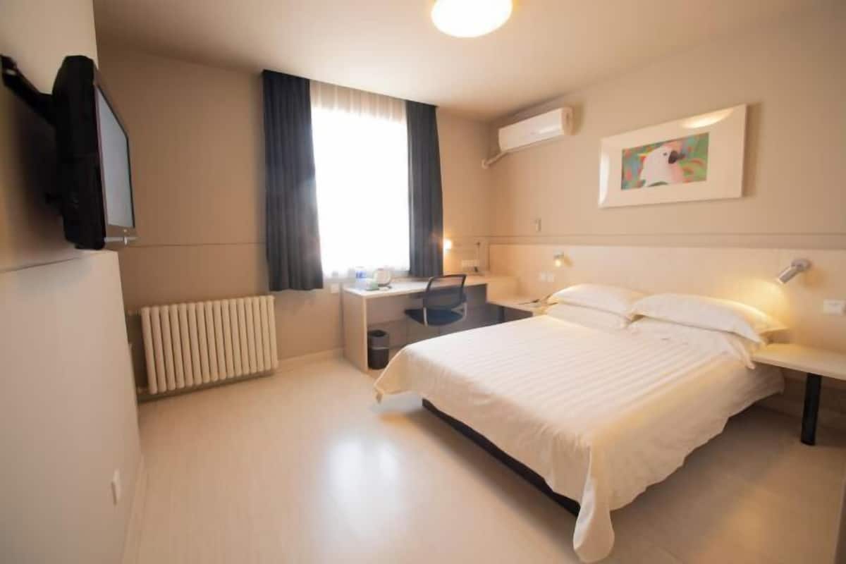 A hotel room with a bed, a desk, a chair, a radiator, a window with curtains, and a wall-mounted air conditioner.