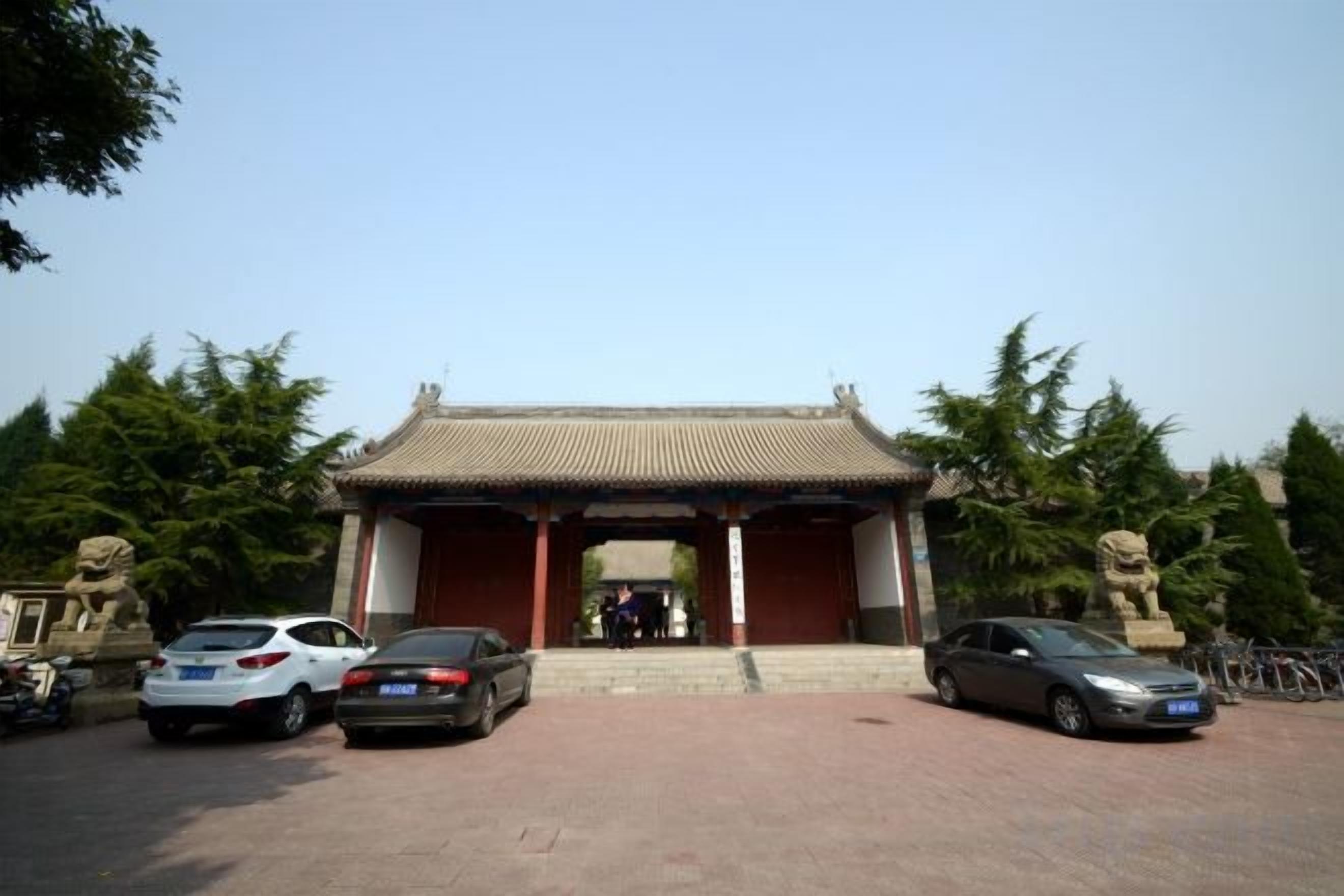 A traditional building with a tiled roof and a central entrance. There are cars parked in front, and a statue is visible to the right.