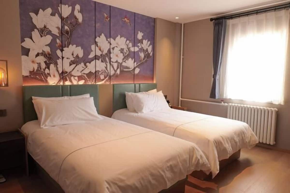 A hotel room with two beds, a large floral artwork on the wall, a window with curtains, and a radiator.