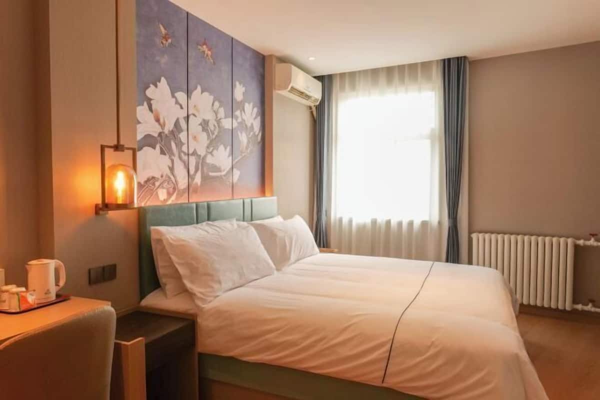 A hotel room with a bed, a desk with a chair, a painting on the wall, and a window with curtains.