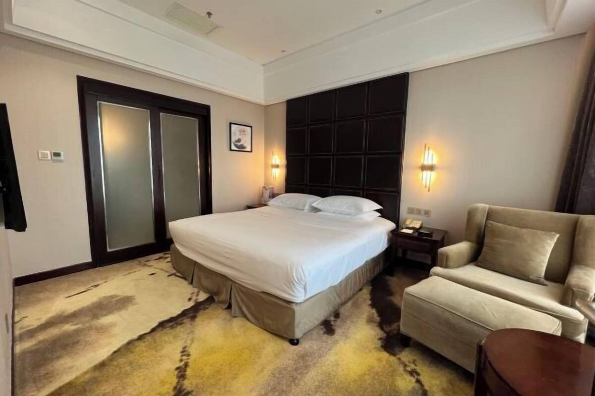 Habitación de hotel con una cama grande, un sofá, una mesita de noche y una lámpara fijada a la pared.