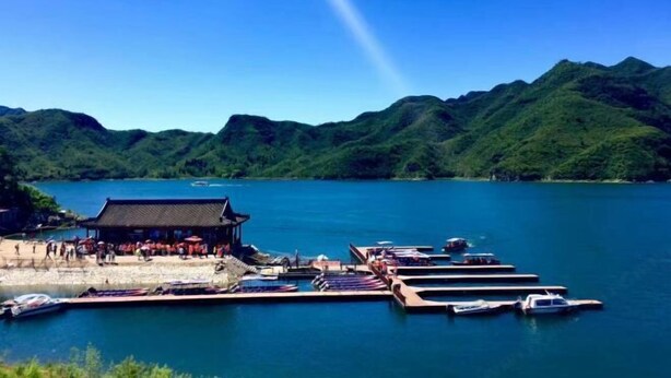 A lakeside dock with boats, a traditional building, and lush green hills in the background. - Zhuozheng International Hotel (Baoding)
