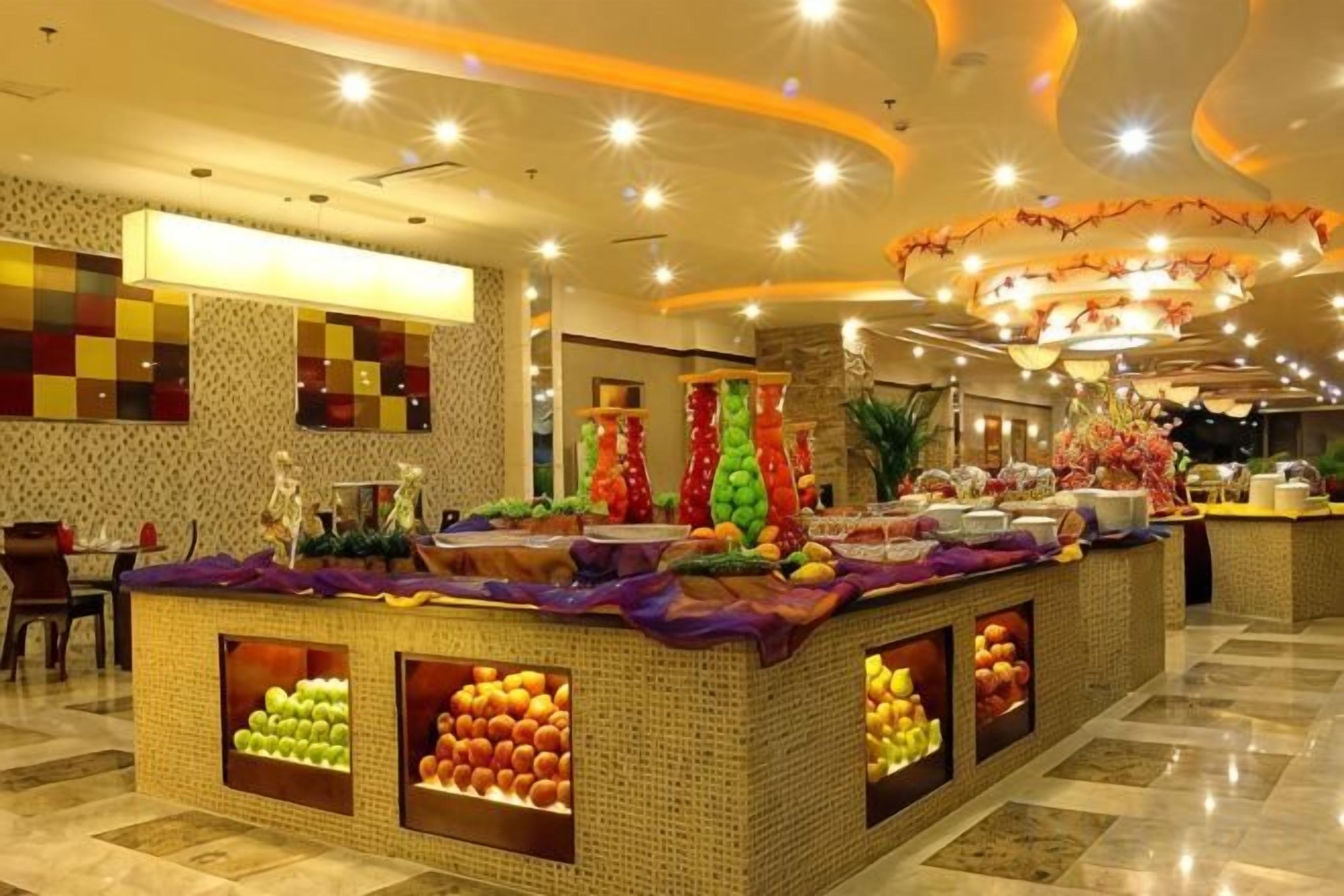 Un vestíbulo de hotel con una zona de buffet central que presenta exhibiciones de frutas y un área de descanso con mesas y sillas.