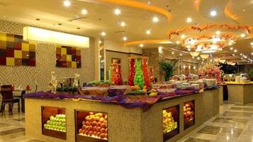 Un vestíbulo de hotel con una zona de buffet central que presenta exhibiciones de frutas y un área de descanso con mesas y sillas.