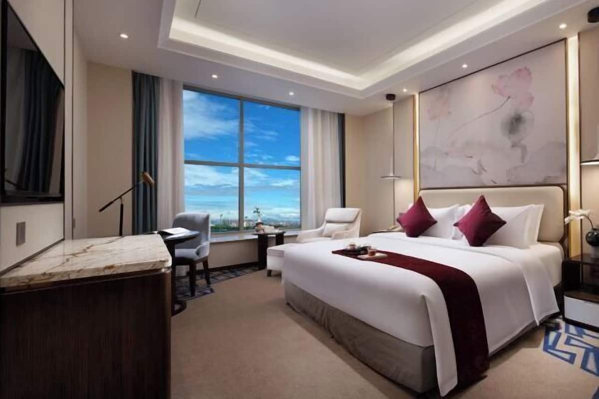 Habitación de hotel con una cama grande, un escritorio, una silla y vista al cielo a través de un ventanal.