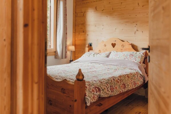 A wooden bed with a floral bedspread, a nightstand with a lamp, and a window with curtains.