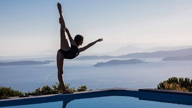 Eine Person vollführt eine Yoga-Pose am Poolrand mit Blick auf Berge und Wasser in der Ferne.