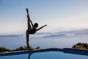 Una persona realizando una postura de yoga en el borde de una piscina, con vistas panorámicas de montañas y agua al fondo.