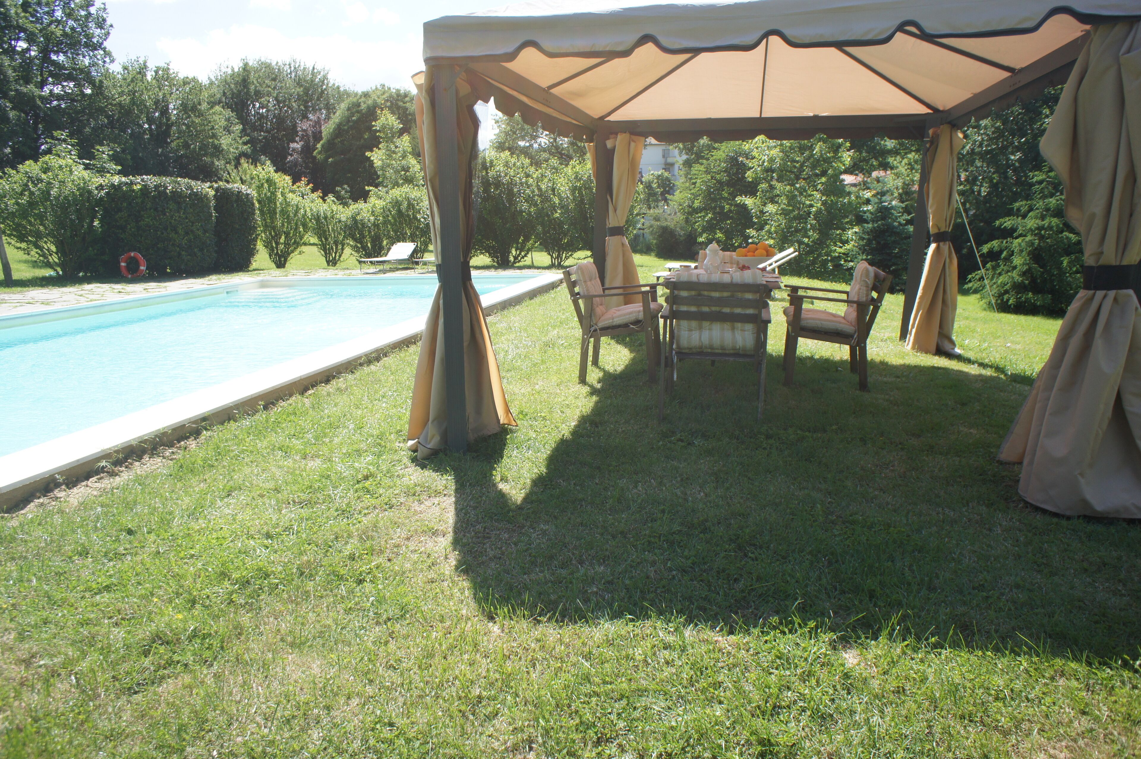 A covered outdoor seating area with a pool, grass, and trees in the background.