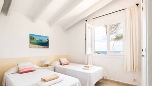 A hotel room with two beds, a painting of a beach, a window with a view of the sea, and a curtain. - Binire · Villa BiniRe ❤️ private pool ❤️ Free AC/Wifi (Binibequer)