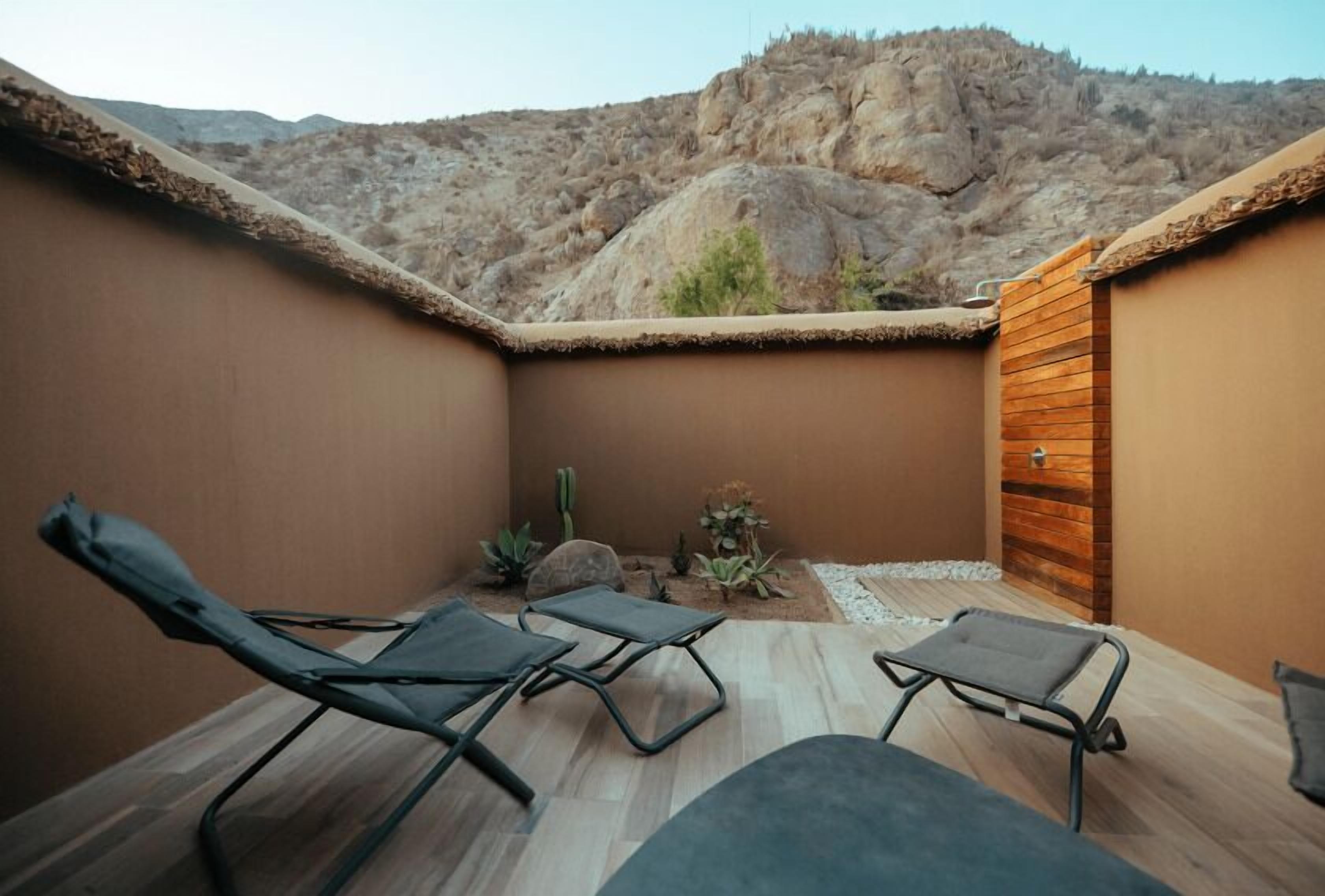 A sunlit patio with lounge chairs, a wooden door, and a view of rocky hills.