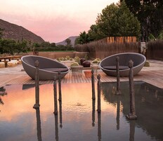 Un espacio al aire libre tranquilo con dos grandes sillones modernos de forma redondeada y una pequeña mesa, rodeado de cactus y plantas de desierto, con un elemento acuático sereno en primer plano.