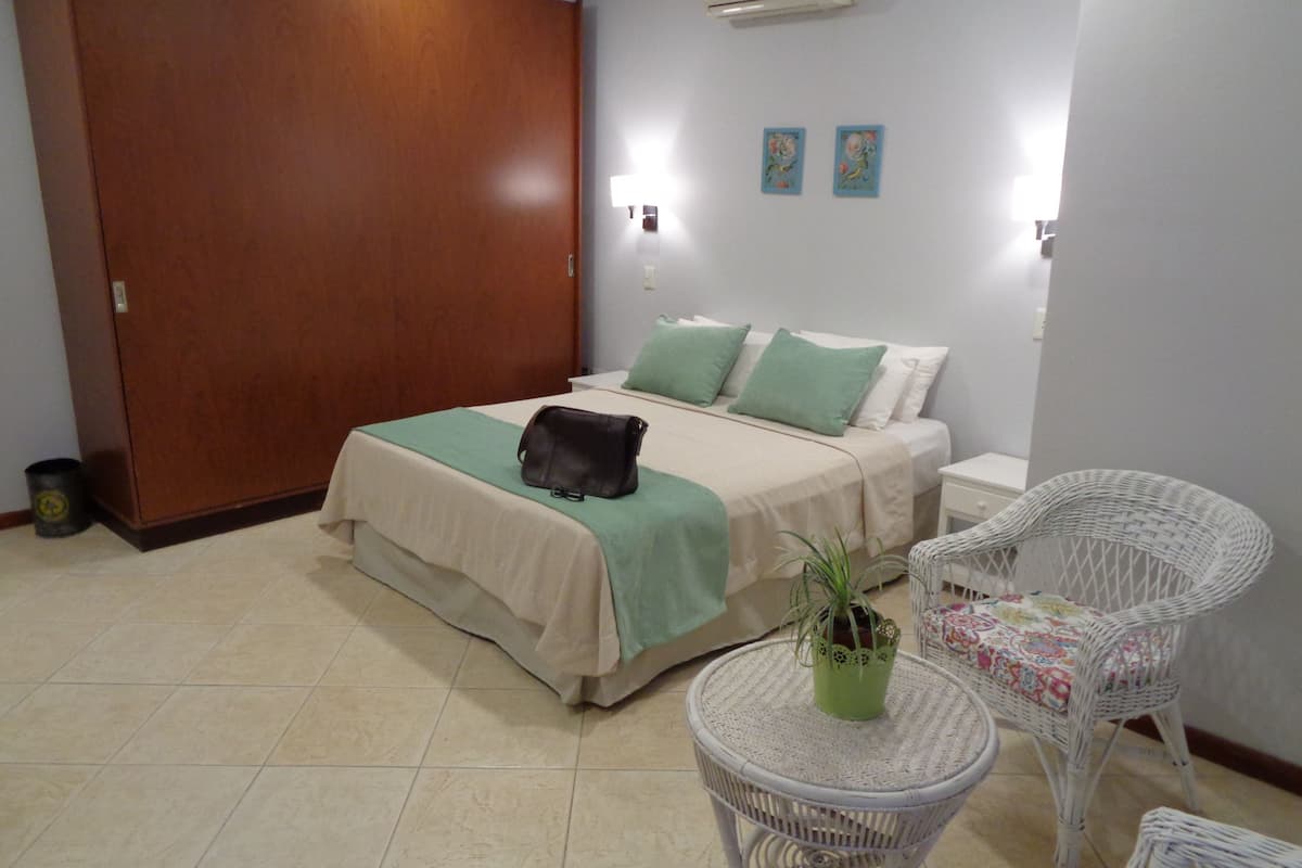 A hotel room with a bed, a small round table with a plant, two white wicker chairs, and a wooden wardrobe.