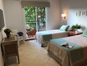 Una camera d'albergo con due letti, una sedia, un tavolino e un balcone con vista sul verde.