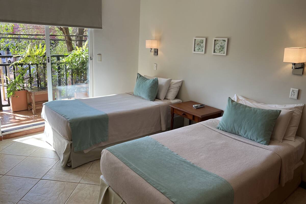 A hotel room with two beds, a small table, and a window with a view of greenery.