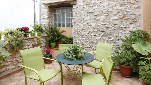 A patio with a round table and four green chairs, surrounded by potted plants and a stone wall.