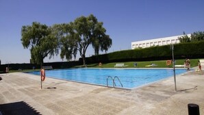 A swimming pool with a clear blue water, surrounded by a paved area with trees and a building in the background.