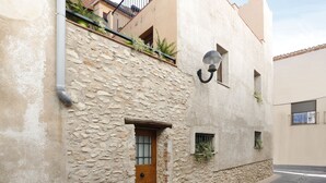 A narrow street with a stone building featuring a wooden door, a small window with plants, and a metal lamp post.