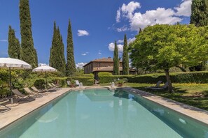 Une piscine entourée de chaises longues, un bâtiment en pierre et de grands arbres en arrière-plan.