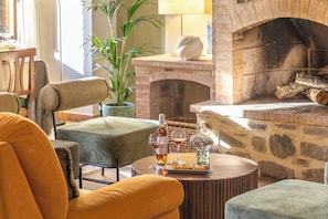 A cozy living room with a stone fireplace, comfortable seating, a table with a bottle and glasses, and a potted plant.