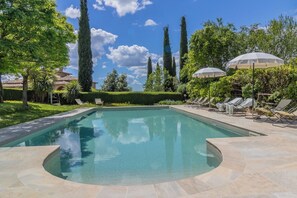 Ein rechteckiger Pool mit geschwungener Kante, umgeben von Liegestühlen und Sonnenschirmen, mit hohen Bäumen im Hintergrund.